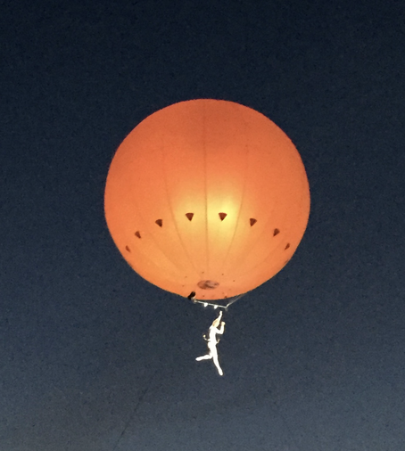 Foto: Nachthimmel, davor ein in Orange angestrahlter Heißluftballon, an dem ein Mensch sacht zum Boden schwebt