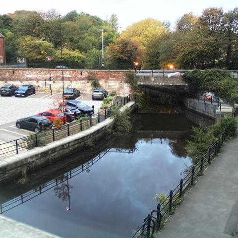 Photograph of the tidal stretch of Ouseburn River at south side of Ouseburn Bridge in Newcastle upon Tyne. To the left (south-west) of the bridge is Cut Bank, and to the right (north-east) is Byker Bank. In the foreground is the pedestrian Riverside Walk and the photo also spans some surrounding built-up areas and trees, with sky in the background. Still image captured from webcam at 07:24 hrs on Saturday 11 October 2025.