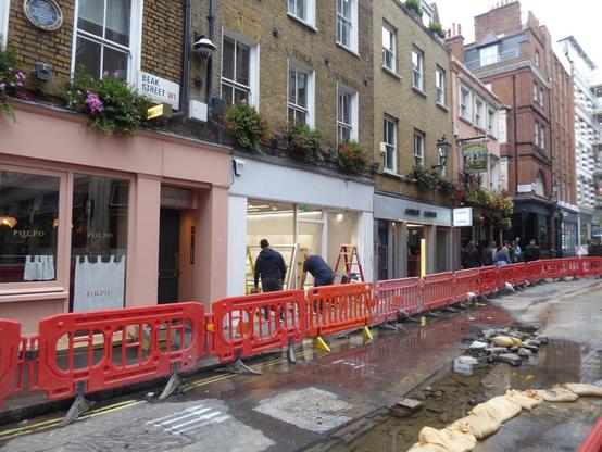 Day. View down street with red barriers along pavement. Centre of road has shallow trench dug in the middle and is filled with water.
