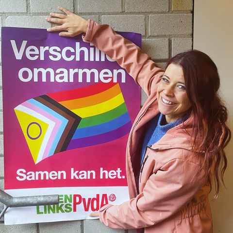 Poster met tekst “verschillen omarmen, samen kan het” met daarin een Pride-vlag en het logo van GroenLinks-PvdA. Poster is van de campagne van twee jaar geleden. Ik hou die vast tegen een muur en sta met een roze jas aan. Ik kijk over mijn schouder de camera in. 