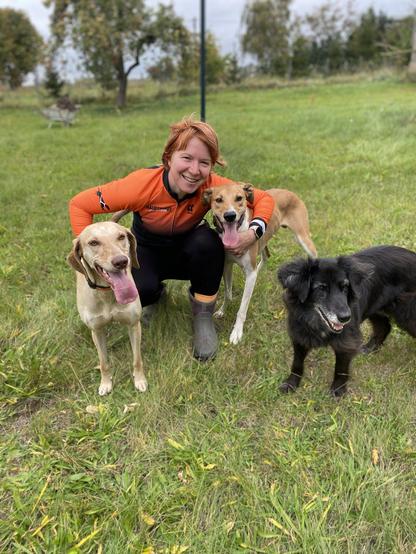 Eine Frau mit schwarz-oranger Sportkleidung kniet auf einer Wiese. Links hat sie einen braun-weißen, rechts einen blonden Hund im Arm. Ein schwarzer, älterer Hund steht auch noch im Bild.
