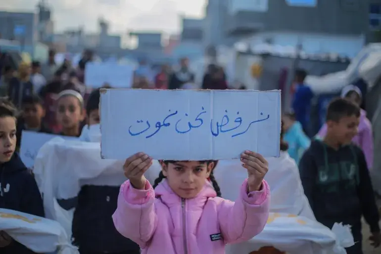 A young girl in pink jacket holds a sign in Arabic saying "We refuse to die."