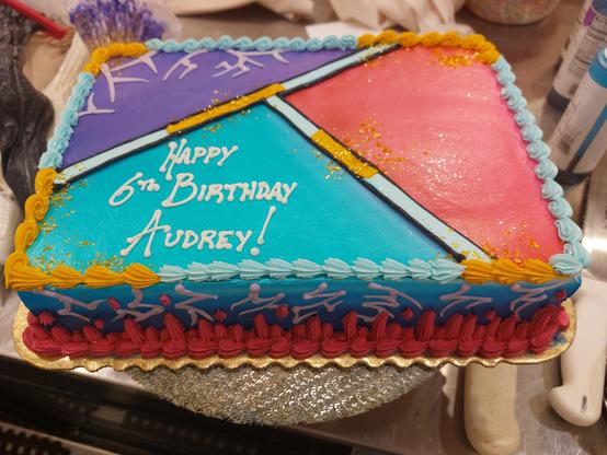 A sheet cake decorated with a KPOP Demon Hunters theme. The top is iced in 3 colors, each representing one of the trio; red/magenta for Mira, aqua/teal for Zoey, and purple (with some demon patterns at the edge) for Rumi. The borders are a mixture of light blue and gold, similar to the semi-charged Honmoon. The sides of the cake are sprayed blue, which darkens as it approaches the bottom of the cake. The top of each of the sides is a sequence of demon patterns, while the lower half is covered in dark pink flames to represent Gwi Ma.

Printed in white icing, in the teal Zoey section, is "Happy 6th Birthday Audrey!"