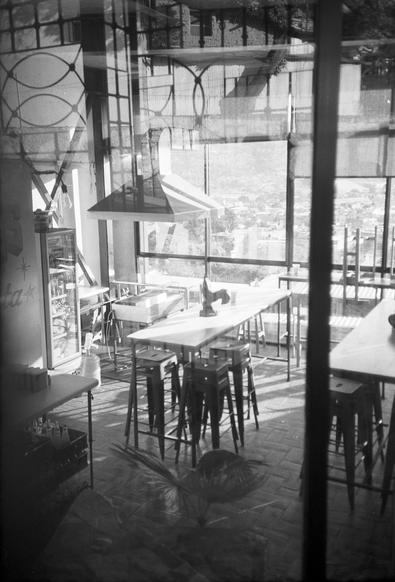 Photo N&B de la salle ensoleillée d'un petit restaurant en surplomb de la ville de Oaxaca. Le restaurant est fermé, la salle est vide, on distingue des reflets de la rue à travers la vitre devant laquelle fût prise la photo.