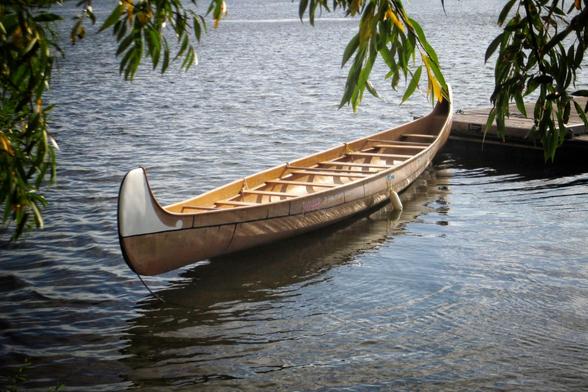 A large multi person canoe sits tied up to  a dock. The canoe is tied up at its bow and has floated around pointing away from the dock. Branches from nearby trees hang down in front of the picture.