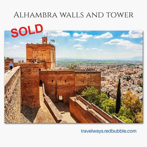 The ancient stone walls towering over the Alhambra Palace and Moorish architectural complex overlook the city of Granada, Spain under a bright blue summer sky. Visitors walk along the fortress, enjoying panoramic views of the landscape and the distant horizon. 