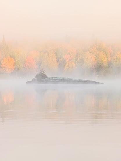 A misty lake at dawn with an island and vibrant autumn trees in the background.
