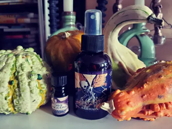 A 5ml perfume oil bottle of Dark Macadamia, and a bottle of Samhain home & linen spray from Black Phoenix Alchemy Lab, surrounded by various orange & green gourds.