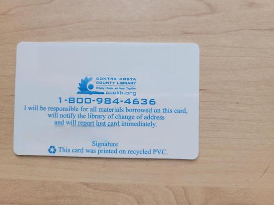 Photo of the back of a library card on a maple desk surface. The card design is white with blue text, showing the same text as the front, but with the phone number (18009844636) and pledge: "I will be responsible for al materials borrowed on this card, will notify the library of change of address and will report lost card immediately." The bar code and signature have been digitally erased. Footer text reads "This card was printed on recycled PVC"
