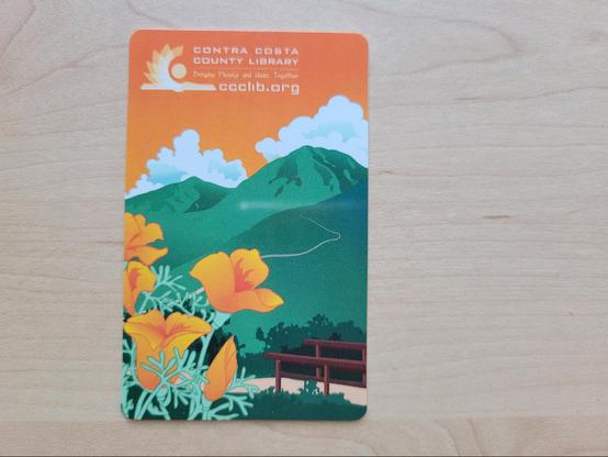 Photo of a library card on a maple desk surface. The card design shows an illustration of golden poppies in the foreground, with a trail leading up into the green mountains in the far distance. The sky is orange with white puffy clouds. The text in the top-left reads "Contra Costa County Library", "Bringing People and Ideas Together" and "ccclib.org"