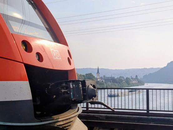 A fotografia mostra a parte da frente de um trem vermelho de perto. A frente do trem é angular, com uma grande janela inclinada, e o logotipo "DB" está visível. O trem está em uma linha férrea com uma cerca de metal ao lado, e há um corpo de água, paisagem e colinas ao fundo.