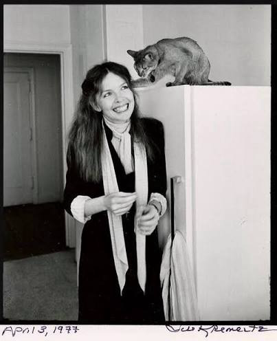 Actress Diane Keaton, wearing a dark coat and light scarf, smiling and standing next to a refrigerator upon which a tabby cat is perched. 