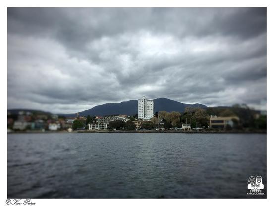 A photograph showing the approach to Hobart, Tasmania, with a focus on a tall, white building in the center. 
The foreground is a dark, slightly blurred body of water (the Derwent River).

The mid ground features the shoreline with low rise buildings and trees, and the background is dominated by the dark, majestic outline of Mount Wellington under a dramatic, cloudy sky.

The image has a tilt-shift effect, blurring the top and bottom.