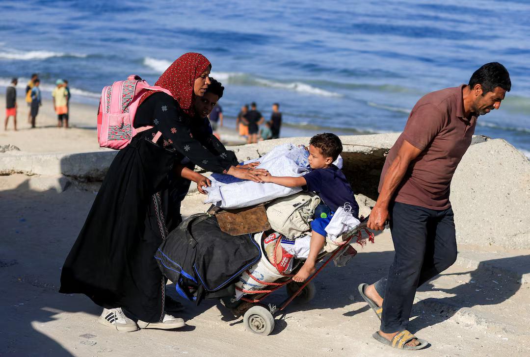 A father is pulling a luggage trolley with all their belongings, the mother is reassuring their young son sitting on top, along the beach road.