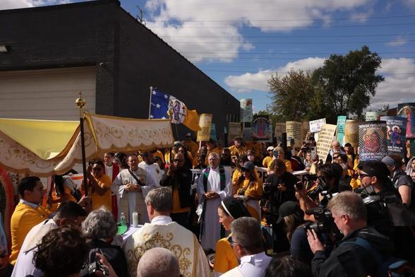 A large group of people participate in a religious protest, holding signs and banners, some wearing yellow robes, under a partly cloudy sky at the Chicago ICE facility in the precoession of the Eucharist.