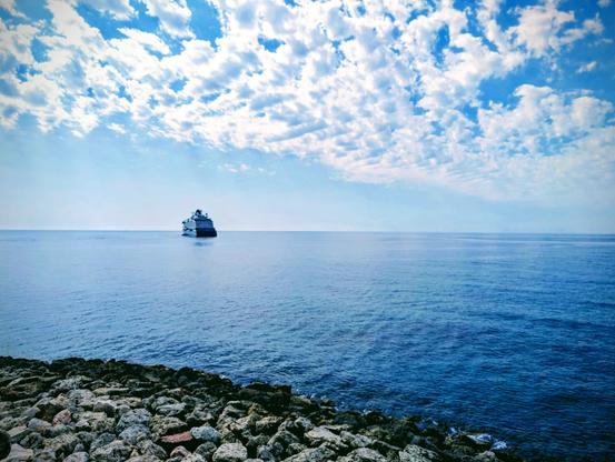 On a calm blue Mediterranean sea beneath a sky dappled with white clouds, a cruise ship is departing from Pafos. In the foreground gentle waves lap against a breakwater of boulders 