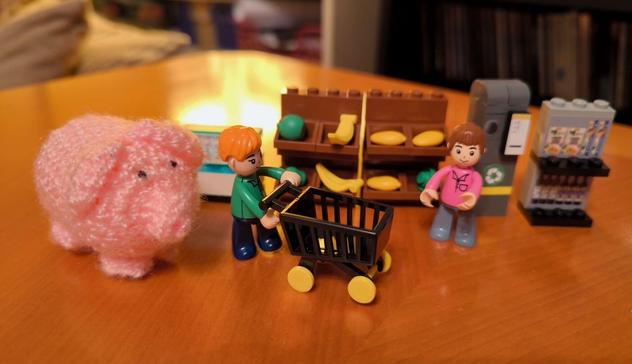Ein haekelschwein und zwei Klemmbausteinfiguren beim Shoppen neben Supermarktregalen und mit einem Einkaufswagen.