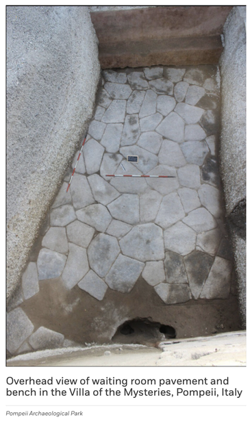 Overhead view of a stone pavement made of irregular light-gray slabs in a rectangular room with rough walls. A low bench runs along the top edge.