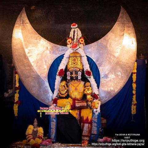Idol of Hindu deity Krishna in yellow (red border around ankles) seated left leg down, right leg folded at the knee, right foot placed over left knee; gold pot in left hand, right hand in blessing posture; wearing gold shoes, gold palm coverings, gold crown, and lots of gold ornaments. A string of white flowers around Krishna. A large silver half moon behind Krishna. A large circular hope within the moon and the void is filled with a matte blue silk cloth.