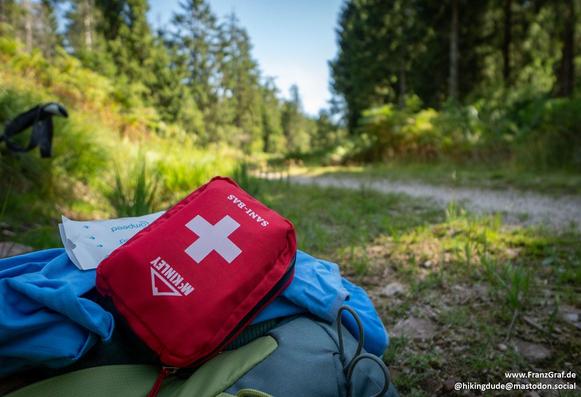 A well-prepared hiking setup featuring a bright red first aid kit prominently placed on top of a backpack, ready for a safe and enjoyable outdoor adventure. The backpack rests on a grassy forest floor, with a scenic trail and lush green trees in the background, creating a sense of readiness and excitement for a day of exploration in nature. The image highlights the importance of safety and preparedness while enjoying the beauty of the great outdoors.