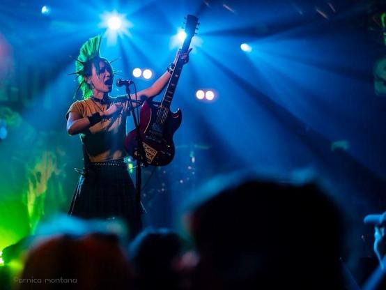 Eine Künstlerin mit grüner Irokesenfrisur spielt auf der Bühne eine E-Gitarre. Die Szene wird von blauen und grünen Lichtern beleuchtet, die eine lebhafte Atmosphäre schaffen. Das Publikum ist teilweise im Vordergrund zu sehen und konzentriert sich auf die energiegeladene Darbietung.
