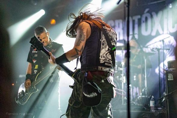 Ein Live-Auftritt, bei dem zwei männliche Musiker auf der Bühne stehen. Der eine spielt Gitarre mit langen Haaren, der andere spielt Bass. Beide sind in dunkler Kleidung mit Bandlogos gekleidet. Helle Bühnenlichter schaffen eine dynamische Atmosphäre im Hintergrund.