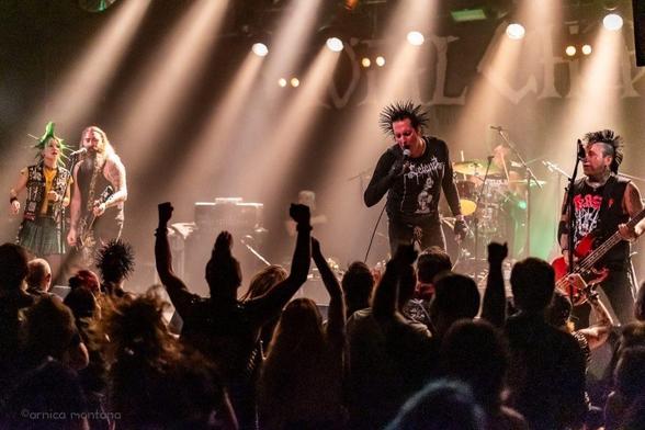 Auf der Bühne tritt eine Punk-Rock-Band auf, die aus mehreren Mitgliedern mit markanten Punk-Frisuren und -Klamotten besteht. Das Publikum ist engagiert und hebt begeistert die Fäuste. Helle Bühnenlichter beleuchten die Musiker und schaffen eine lebendige Atmosphäre.