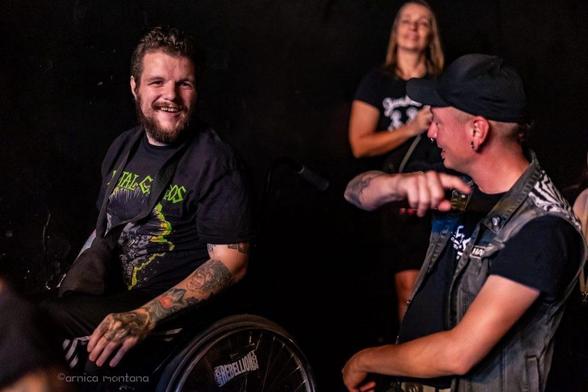 Ein Mann im Rollstuhl, der ein schwarzes T-Shirt mit einem grafischen Muster trägt, lächelt und unterhält sich mit einem anderen Mann, der auf ihn zeigt und eine Weste trägt. Eine Frau im Hintergrund genießt den Moment und trägt zu einer lebhaften Atmosphäre bei. Die Umgebung ist leger.