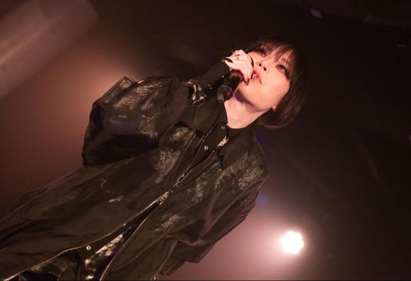 Photo shows singer-songwriter NANO performing on stage. She is wearing black clothing and is holding a microphone close tho her mouth, while the lighting is bright making it a dramatic photo. Photo by JJ Grant (Wonderlens)