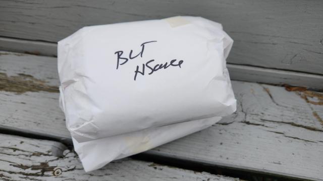 Two sandwiches wrapped in white waxed paper. The top one has writing in sharpie that reads BLT HSauce. The sandwiches are sitting on an outdoor wooden staircase painted Gray.