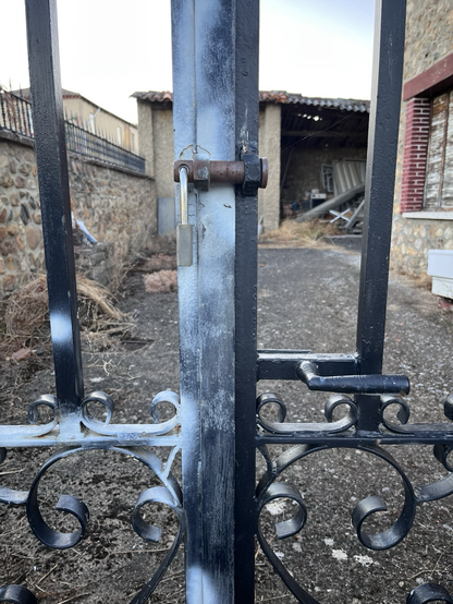 ALT Parte central de una verja exterior, de hierro negro, cerrada con un candado. Por dentro de la propiedad, a la izquierda un muro de piedra, en la zona central camino de entrada y al fondo una construcción de pueblo para almacenaje, con el tejado en mal estado. Tiene una zona de entrada muy amplia, abierta, y dentro vemos un montón de trastos sin valor. 