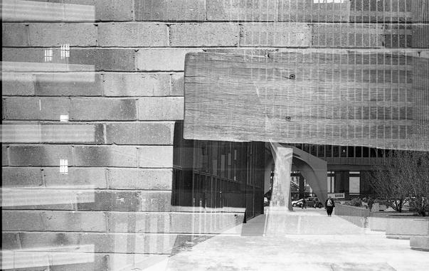 A black-and-white double exposure combining two scenes: a textured brick wall and a large wooden beam overlay an urban walkway. In the second exposure, a modern sculpture and pedestrians appear faintly beneath, blending architecture and texture into a surreal composite.