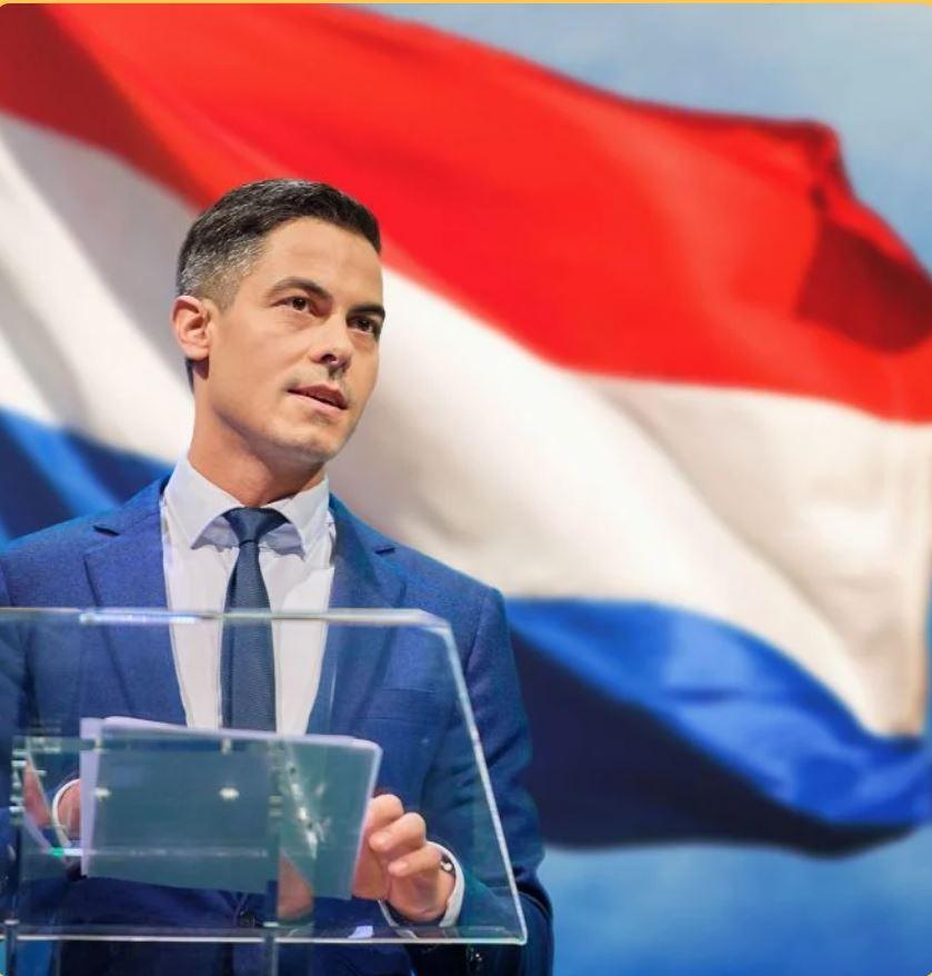 Rob Jetten op een campagnefoto waar Wilders zich niet voot zou schamen. 
Kort geschoren kapsel, jaren 'dertig, met een grote wapperende Nederlandse vlag achter zich, op een katheder van plexiglas waar een stapeltje papieren op liggen.