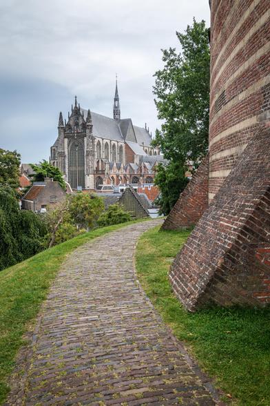 A photograph looking down a cobbled path towards a large, stone, steepled Gothic church. On the right is a banded brick wall, part of a circular fort.
