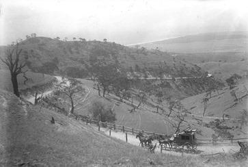 A black and white, outdoor photograph shows a grassy, hilly landscape with a dirt path winding through it. The scene is dominated by a large, gently sloping hill in the foreground, covered in sparse vegetation and trees. A dirt path snakes its way up the hill, with a group of people and what appears to be horse-drawn carriages visible along the path. In the distance, the landscape continues to roll with more hills and a hazy sky. The photograph appears to be taken from a slightly elevated perspective, looking down towards the path and the distant hills.