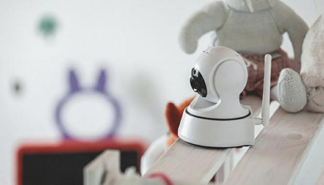 A small, dome-shaped white indoor camera (Nanny Cams) on a shelf in a nursery setting with stuffed animals in the background, illustrating discreet home surveillance for families.

