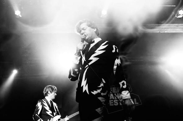 Der Sänger von "the Hives" leicht von unten fotografiert. Er singt in ein mikro und trägt einen Anzug mit grafischem Muster. Im Hintergrund seitlich und nur halb zu sehen ein Gitarrenspieler