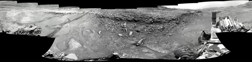 A black and white mosaic of 20 images capturing a Martian landscape, with some elements of the Mars rover Curiosity displayed in the foreground. The tracks of rover descending into the trough can be seen on the left hand side of the mosaic.