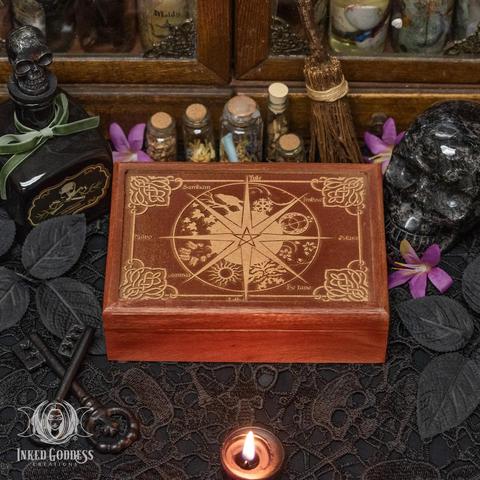 An example of the "Wheel of the Year Wooden Box" product is laid out on a black lace-covered surface. It features a carved design of the Wheel of the Year on its lid, and each sabbat has its own imagery within the Wheel. At the center of the Wheel is a pentacle, and the corners feature Celtic knot-like designs. The box is a medium reddish brown with a lighter finish on the carving. Presented by Inked Goddess Creations.