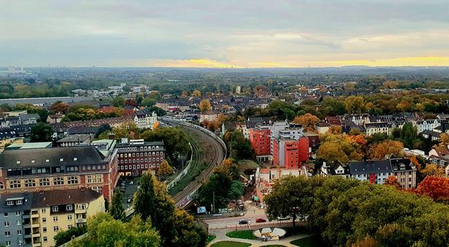 Luftbild zeigt daa Stadtgebiet Bochums bis Duisburg am Horizont. Die Stadt besteht aus vielen Gebäuden in verschiedenen Größen und Farben, die durch Straßen getrennt sind. Ein langer, gewundenes Zuggleis schwingt sich durch die Szene. Bäume sind zwischen den Gebäuden und entlang der Straßen verstreut, und einige Bäume sind in Herbstfarben gekleidet. Am unteren Rand des Bildes ist ein kleiner Park der Wiese vor dem Bergbaumuseum. 