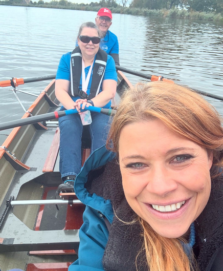 Selfie op het water waarbij ik de camera vasthou en links mijn gezicht lachend in beeld is. Ik draag een blauwe jas met capuchon. Achter mij zit Maureen die twee roeispanen vasthoudt, want we zitten in een boot. Weer achter haar een van de vrijwilligers. Het is op de foto ook te zien dat we op het water zijn.