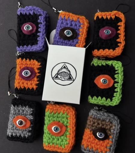 Eight crocheted mini book charms, each with a plastic eye on their cover. In the middle in an open Mega Mystery Art Machine box with its logo in the center.
