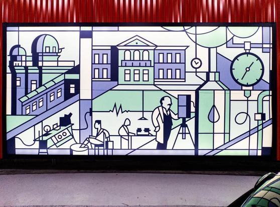 A picture of a back illuminated graphic in a car park, in purple, green, and black, showing some telescopes, scientists, buildings, and a clock. The wall behind is red painted metal and there is a car in the lower right, reflecting some of the mural.