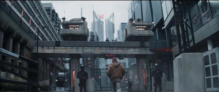 Screenshot from The Running Man trailer. Rear view of man approaching gates under a bridge, flanked by black-armoured security guards. A forbidding cityscape surrounds.