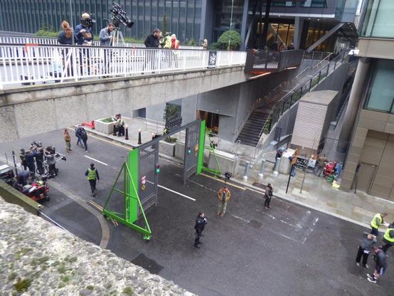 Bridge over a road. Below, gates flanked by fluorescent green panels with three actors in front of them. Various cameras set up for filming. Day.