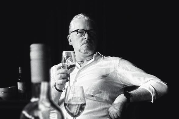 Schwarz-Weiß-Bild eines sitzenden Mannes mit weißem Hemd, getrimmtem Bart und schwarzer Brille. Er stützt sich mit einem Arm auf der Stuhllehne ab, hält ein geknicktes Glas Wein in seiner rechten Hand und schaut ernst in die Kamera. 