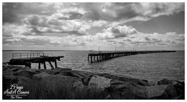 A black and white photograph of the historic, partially dilapidated Tanker Jetty in Esperance, Western Australia.

The remains of the jetty stretch out into the ocean under a dramatic, cloudy sky. In the foreground, large rocks and some coastal grass frame the image.