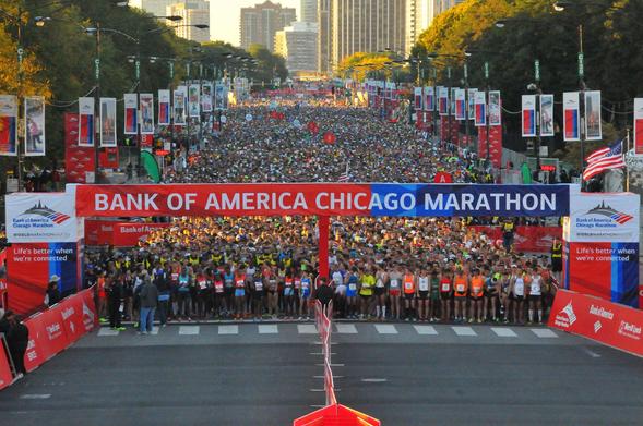 Thousands of runners fill the streets of downtown Chicago at the start of the Bank of America Chicago Marathon. A large red and blue banner marks the starting line, surrounded by cheering crowds, colorful race bibs, and city skyscrapers in the background under clear skies.
