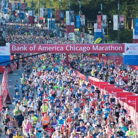 Thousands of runners fill the streets of downtown Chicago at the start of the Bank of America Chicago Marathon. A large red and blue banner marks the starting line, surrounded by cheering crowds, colorful race bibs, and city skyscrapers in the background under clear skies.