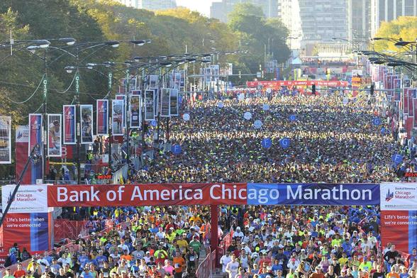 Thousands of runners fill the streets of downtown Chicago at the start of the Bank of America Chicago Marathon. A large red and blue banner marks the starting line, surrounded by cheering crowds, colorful race bibs, and city skyscrapers in the background under clear skies.